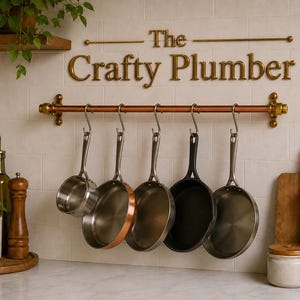 May include: A copper and brass pot rack with six hanging pans. The Crafty Plumber is displayed above the rack. The pans are stainless steel and copper. A wooden shelf with a plant is above the counter.