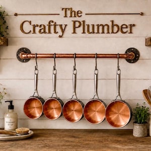 May include: A copper pipe wall rack with five hanging copper-bottomed saucepans featuring silver handles. The wall displays the text The Crafty Plumber in gold lettering above the rack, flanked by wooden shelves with plants and a utensil crock.