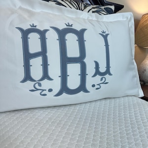 Peut inclure: Taie d'oreiller blanche avec un grand monogramme gris des lettres RBJ. Le monogramme est dans une police décorative avec des détails de couronne. La taie d'oreiller est sur un lit avec un couvre-lit blanc matelassé.