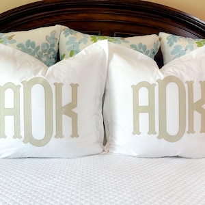 Peut inclure: Deux oreillers décoratifs blancs avec un monogramme beige clair. Les oreillers sont posés sur un lit blanc, avec une tête de lit marron foncé en arrière-plan. Des oreillers à motifs floraux sont également visibles.