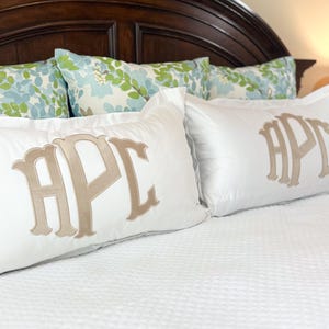 Op de afbeelding: Twee witte rechthoekige kussens met geborduurde initialen in een lichtbruine kleur. De kussens liggen op een wit gewatteerd sprei, met kussens met bloemenpatroon en een donkerbruin hoofdbord op de achtergrond. De initialen zijn "HPC".