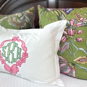 Op de afbeelding: Witte kussensloop met een roze en groen monogram, omlijst door een roze bloemenkrans en strik. Twee sierkussens met bloempatronen in groen en roze bevinden zich achter de kussensloop. De kussens liggen op een wit bed.