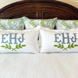 May include: White rectangular pillowcases with blue embroidered initials and green leaf accents. The pillows are on a bed with a white quilted bedspread and floral patterned pillows.