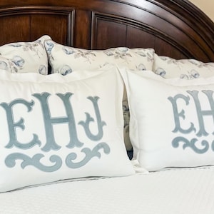 Peut inclure: Deux taies d'oreiller blanches avec les lettres "EHJ" brodées en bleu clair. Les oreillers sont sur un lit blanc avec une tête de lit en bois brun foncé en arrière-plan. Des oreillers décoratifs sont également visibles.