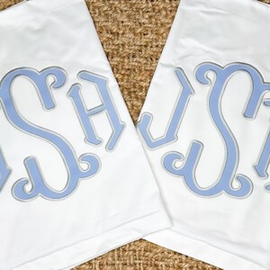 Könnte beinhalten: Zwei weiße Küchentücher mit einem hellblauen Monogramm der Buchstaben JSH. Das Monogramm ist in einer traditionellen Schriftart.
