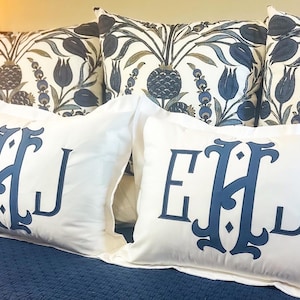 Könnte beinhalten: Weiße Kissenbezüge mit einem marineblauen EHJ-Monogramm. Die Kissen liegen auf einer marineblauen Tagesdecke. Dekorative Kissen mit einem floralen Muster in Marineblau und Weiß sind im Hintergrund zu sehen.