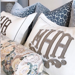 May include: A white pillow with the monogram "MHA" embroidered in brown. The pillow is on a bed with a floral patterned bolster pillow and blue patterned pillows.