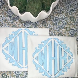 Op de afbeelding: Twee witte kussenslopen met een lichtblauw monogramontwerp. Het monogram toont de letters "WHK" in een decoratieve, sierlijke stijl, geplaatst in een lichtblauw kader. De kussenslopen liggen op een stof met patroon.