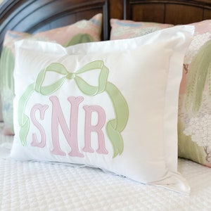 Op de afbeelding: Een wit kussen met een groene strik en roze geborduurde initialen "SNR" op een wit bed.