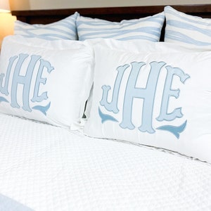 May include: Two white rectangular pillows embroidered with light blue initials JHE. The pillows rest on a white bed with a quilted pattern. Two striped blue and white pillows are in the background.