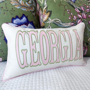 May include: A rectangular white throw pillow featuring the name GEORGIA embroidered in large, light pink letters with a sage green outline. The pillow has matching light pink piping along the edges and a smooth, solid white fabric finish.
