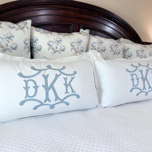 Peut inclure: Deux taies d'oreiller rectangulaires blanches avec un monogramme bleu clair des lettres DKK. Le monogramme est dans une police décorative et ornée. Les taies d'oreiller sont sur un couvre-lit blanc, avec des oreillers à motifs floraux en arrière-plan.