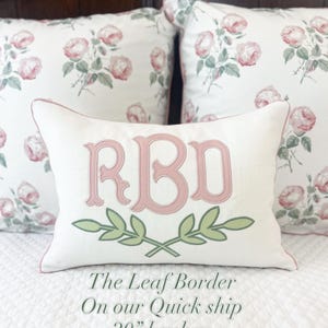 Könnte beinhalten: Ein rechteckiges Lendenkissen mit weißem Stoffbezug, das ein in Rosa gesticktes Monogramm RBD über einem in Grün gestickten Blattzweig-Design zeigt. Das Kissen hat eine rosa Paspelierung an den Rändern und ist vor zwei quadratischen Kissen mit rosa Rosenmustern platziert.