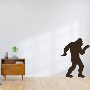 May include: A black silhouette of Bigfoot walking with one arm raised, as if waving. The silhouette is on a white wall.