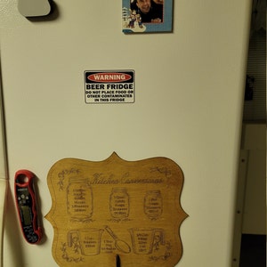 May include: A wooden sign with the text "Kitchen Conversions" and a list of measurements for different units, including gallons, quarts, pints, cups, ounces, and milliliters. The sign is attached to a refrigerator with a key hook.