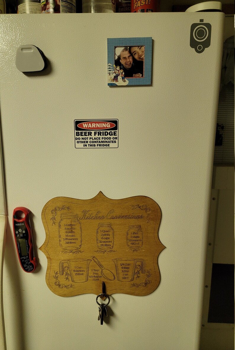 May include: A wooden sign with the text "Kitchen Conversions" and a list of measurements for different units, including gallons, quarts, pints, cups, ounces, and milliliters. The sign is attached to a refrigerator with a key hook.
