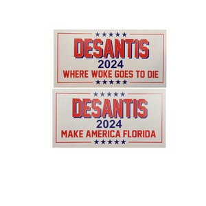 May include: Two white signs with red and blue text. The top sign reads "DESANTIS 2024 WHERE WOKE GOES TO DIE". The bottom sign reads "DESANTIS 2024 MAKE AMERICA FLORIDA".
