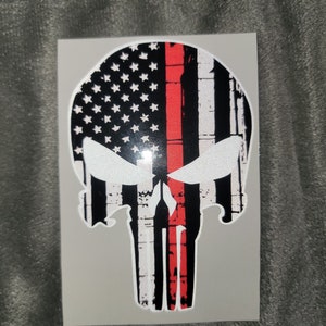 May include: A black and white skull sticker with a red line down the center. The skull is designed to look like the American flag with a thin red line.