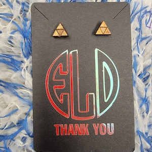 Puede incluir: Un par de pendientes de madera con un diseño triangular. Los pendientes están en una tarjeta negra con el texto "Gracias" impreso en ella.