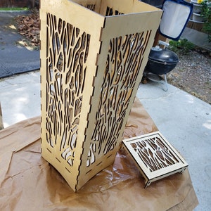 May include: A wooden lampshade with a tree branch design. The lampshade is made of thin plywood and has a square base. The lampshade is sitting on a brown paper surface.