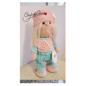 Peut inclure: Une poupée en crochet portant un chapeau rose, un pull bleu clair et un pantalon, et des chaussures blanches. La poupée a les cheveux blonds et un sac à main rose et blanc.