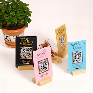 May include: Four business cards with QR codes are displayed on wooden stands. The cards are black, pink, blue, and gold. The text on the cards includes "Total Elegance Candle Company", "EverGray Artistry", "Leave Us A Review", and "Mua Cay".