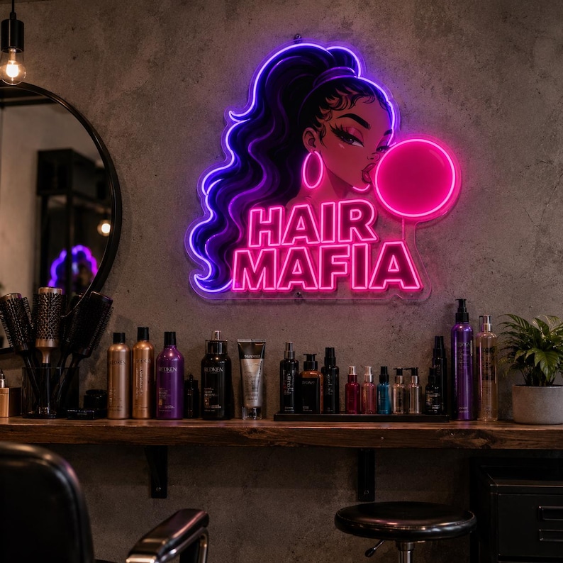 May include: A neon sign featuring a stylized illustration of a person with long dark hair and hoop earrings blowing a pink bubble, with the text HAIR MAFIA in bright pink neon letters below the figure, mounted on a textured gray wall.