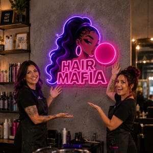 May include: A neon sign featuring a stylized illustration of a person with long dark hair blowing a pink bubble, with the text HAIR MAFIA in bright pink neon letters. The sign is mounted on a gray textured wall, glowing with purple and pink light.