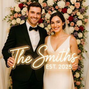 May include: A custom LED neon sign featuring the text The Smiths and EST. 2025 in a warm glowing script font. The sign is mounted on a clear acrylic backing and is held by two people dressed in formal wedding attire in front of a floral backdrop.