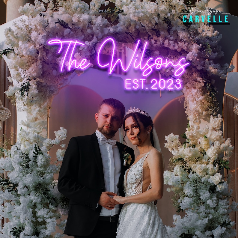 May include: A bright purple neon sign displaying the text The Wilsons and EST. 2023 in a cursive and block font. The sign is mounted above a wedding arch decorated with white flowers, positioned behind a couple in formal wedding attire.