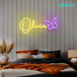May include: A custom neon sign displaying the name Olivia in a bright yellow cursive script, accompanied by a purple neon butterfly outline. The sign is mounted on a vertical paneled wall above a bed with orange and dark gray bedding.