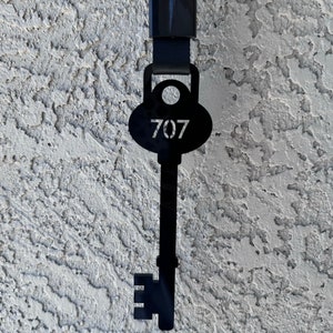 May include: Black metal key chain with a key shaped like a keyhole and the number 707 on the top. The key chain is hanging from a black strap.