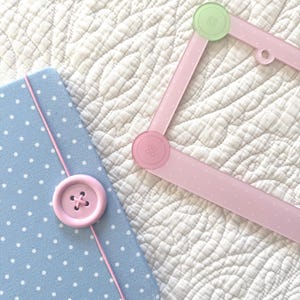 May include: A light blue notebook with white polka dots, secured by a pink elastic band and a large pink button. Beside it lies a pink rectangular frame featuring white polka dots, accented with a light green button and a pink button at corners.