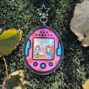 May include: Pink and blue Tamagotchi keychain with a clear plastic cover. The screen shows two cartoon characters in a room with a table and chairs. The keychain has a star-shaped charm.