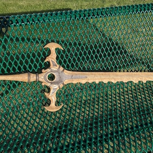 May include: A large, ornate, gold-colored sword with a black blade resting on a green metal bench. The sword has a detailed design with intricate carvings and a pointed tip.