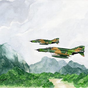 May include: A watercolour painting depicting two camouflage fighter jets flying over a mountain range. The jets are flying in formation, with the leading jet slightly ahead of the trailing jet. The mountains are covered in green foliage, and the sky is a light blue with white clouds.