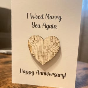 Pode incluir: Um cartão branco com o texto "I Wood Marry You Again" e um coração de madeira com padrão de grão de madeira. O cartão também diz "Happy Anniversary!"