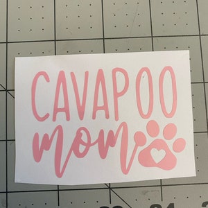 Puede incluir: Calcomanía de vinilo rosa con el texto "CAVAPOO MOM" y una huella de pata rosa con un corazón blanco en su interior.