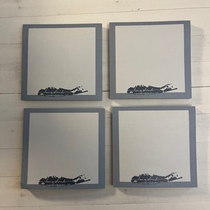 May include: Four square Long Island themed coasters. Each coaster has a white center with a gray border. A black graphic of Long Island and the word "Long Island" is printed on the white center of each coaster.