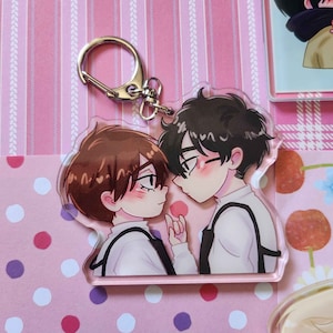 May include: A clear acrylic keychain featuring an anime-style illustration of two male figures with blushing cheeks. The figures are close together, with one holding a small pink object. The keychain has a silver clasp.