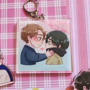 May include: A clear acrylic keychain featuring an illustration of two cartoon figures in an embrace. One figure is wearing a blue suit, and the other is wearing a tan jacket and black scarf. A pink heart floats between them, with a silver keyring at the top.