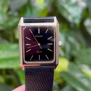 May include: A rectangular gold-toned watch with a black face and a black leather band. The watch face has gold-toned hands and markers, with the brand name "Chander" and the word "Quartz" printed on the face. The watch is set against a blurred green background.