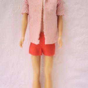 Puede incluir: Una muñeca vintage con el pelo corto castaño, ojos azules y una camisa a rayas rojas y blancas. La muñeca lleva pantalones cortos rojos y está de pie sobre un fondo blanco.