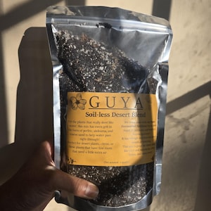 May include: A clear plastic bag of GUYA Soil-less Desert Blend, held by a hand. The bag has a yellow label with text describing the soil mix for plants that don't like water. The mix contains perlite, akdeama, and coarse sand. Net amount: 2 quarts.