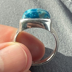 Puede incluir: Un anillo de plata con una piedra preciosa azul engastada en un bisel cuadrado. El anillo se lleva en un dedo.