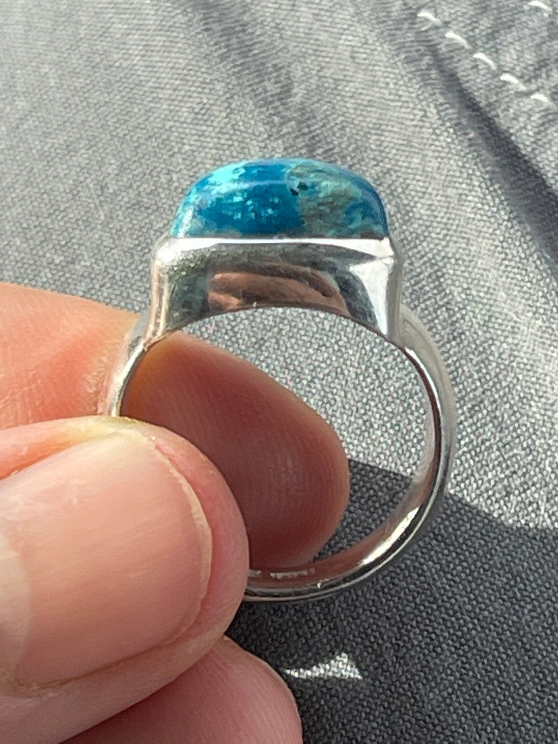 Puede incluir: Un anillo de plata con una piedra preciosa azul engastada en un bisel cuadrado. El anillo se lleva en un dedo.