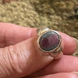 May include: A silver ring featuring a large, oval-shaped gemstone with a deep red and gray color. The ring has a textured band and a bezel setting that holds the stone securely. The ring is a piece of jewelry.
