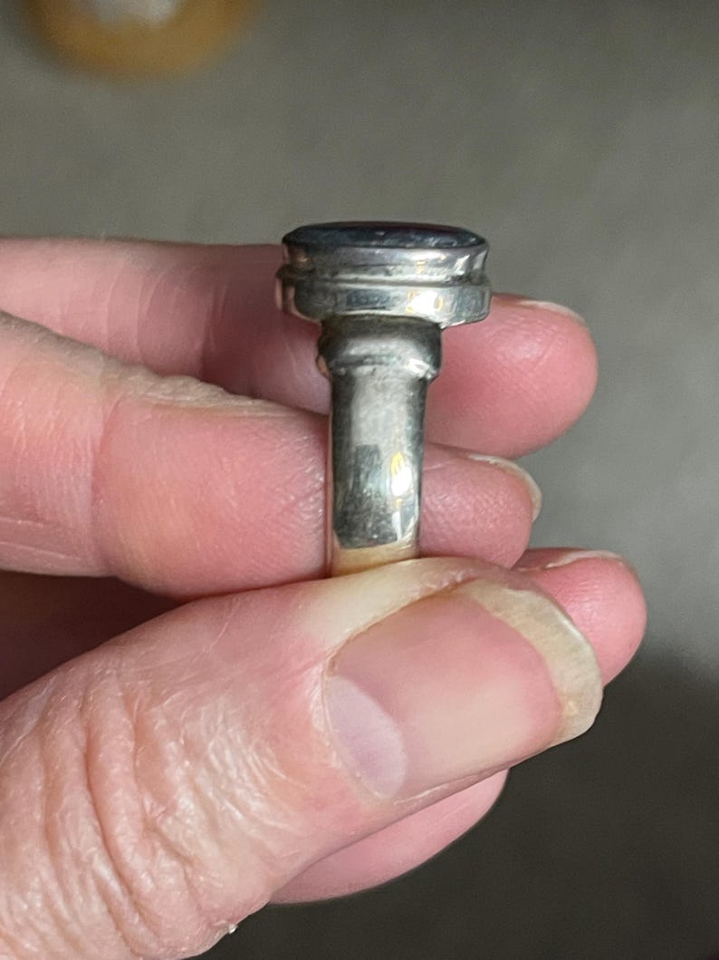 May include: A silver ring with a large, oval-shaped, dark-colored stone. The ring has a thick band and a decorative setting around the stone. The ring is held up by a person's fingers.