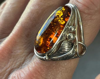 May include: A silver ring featuring a large, oval amber stone with golden inclusions. The ring has a detailed, leaf-like design on the sides and around the amber. The amber is a warm, translucent orange color.