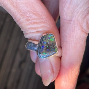 May include: A silver ring with a dark green, blue, and black opal gemstone. The opal is set in a silver band and has a unique, irregular shape.